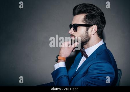 Wunderschön und hübsch. Nahaufnahme eines jungen, gutaussehenden Mannes im Anzug und in der Fliege, der Hand am Kinn hält und wegblickt, während er auf grauem B sitzt Stockfoto