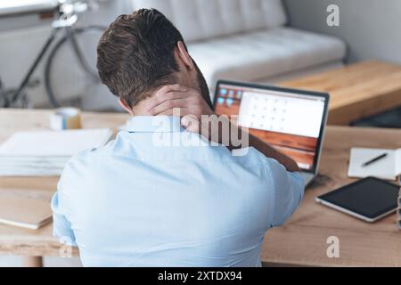 Erschöpft. Rückansicht eines frustrierten jungen Mannes, der erschöpft aussieht und seinen Nacken massiert, während er an seinem Arbeitsplatz sitzt Stockfoto