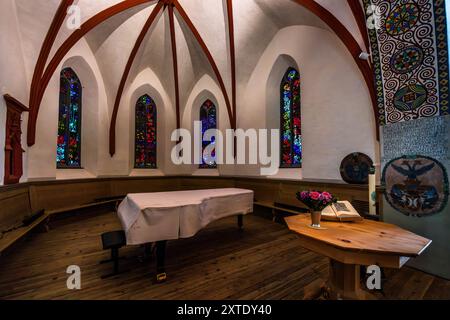 Die Reformierte Kirche St. Johann in Davos hat vier hohe Chorfenster zum Thema Paradies, entworfen von Antonio Augusto Giacometti. Rote Holzrippen stützen das Gewölbe über gekalkten Wänden. Ein Flügel, mit einem weißen Tuch bedeckt, steht vor den Fenstern. Rechts liegen ein hölzerner achteckiger Tisch mit einem Blumenstrauß und einem aufgeschlagenen Buch, ein rundes Wandgemälde und ein Wappen mit einem Adler. Berglistutz, Davos, Graubünden, Schweiz. Stockfoto