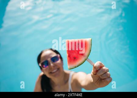 Eine lächelnde Frau, die ein Stück Wassermelone auf einem Stock hält und einen sonnigen Tag am Pool genießt Stockfoto