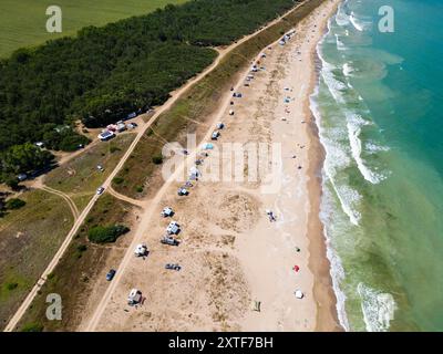 Aus der Vogelperspektive auf einen Sandstrand mit verstreuten Zelten und Sonnenschirmen entlang der Küste. Wellen waschen sanft an die Küste und fangen ein friedliches und Stockfoto