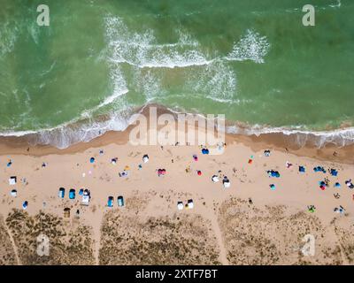 Aus der Vogelperspektive auf einen Sandstrand mit verstreuten Zelten und Sonnenschirmen entlang der Küste. Wellen waschen sanft an die Küste und fangen ein friedliches und Stockfoto