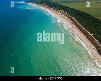 Aus der Vogelperspektive auf einen Sandstrand mit verstreuten Zelten und Sonnenschirmen entlang der Küste. Wellen waschen sanft an die Küste und fangen ein friedliches und Stockfoto