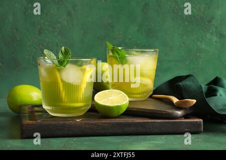 Holztablett mit Gläsern leckerer Limonade, Limetten und Minze auf grünem Grunge Hintergrund Stockfoto