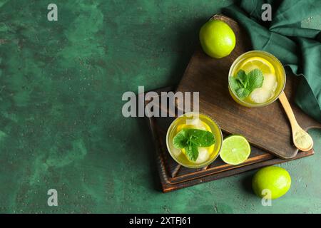 Holztablett mit Gläsern leckerer Limonade, Limetten und Minze auf grünem Grunge Hintergrund Stockfoto