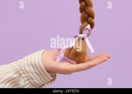 Weibliche Hand mit geflochtenem rotem Haar für Spende und Bewusstseinsband auf violettem Hintergrund Stockfoto