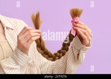 Weibliche Hände mit geflochtenem rotem Haar für Spende und rosa Bewusstseinsband auf lila Hintergrund Stockfoto