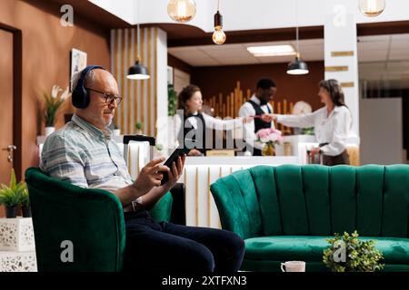 Weibliche Kunden checken an der Hotelrezeption ein, während ältere männliche Reisende auf dem Tablet ihre Zimmer buchen. Pensionierter Seniorenmann mit Kopfhörern, der sein Gerät für den Online-Check-in benutzt. Stockfoto