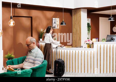 Ältere Reisende warten auf den Check-in an der Hotelrezeption im Loungebereich. Ein älterer Mann liest auf der Couch, während eine ältere Frau mit Koffer an der Kasse wartet. Stockfoto