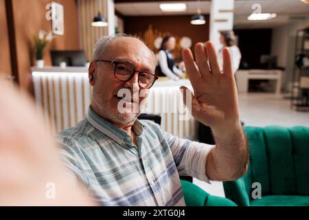 Ein lächelnder älterer Mann in der Hotellounge macht ein Selfie auf seinem Handy, während das Personal anderen Gästen im Hintergrund hilft. Pensionierter Seniorenreisender, der Spaß am Urlaub auf dem Smartphone festnimmt. Stockfoto