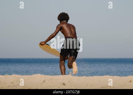 Ein junger Mann, der mit einem Skimboard in der Hand am Strand läuft und bereit ist, die Wellen zu surfen Stockfoto