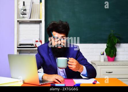 Ein ermüdeter Schullehrer sitzt am Schreibtisch und macht eine Kaffeepause. Verschlafener bärtiger Lehrer trinkt Kaffee im Klassenzimmer. Überarbeiteter Hochschulstuden Stockfoto