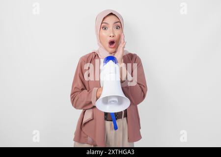 Schockierte asiatische muslimische Frau im Hijab, die Megaphon anschreit und überrascht aussieht, isoliert auf weißem Hintergrund. Stockfoto