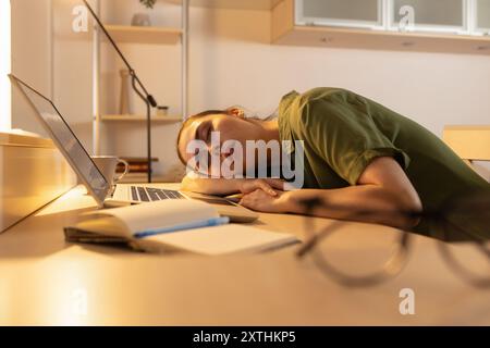 Die junge Frau sitzt mit einem Laptop an ihrem Schreibtisch und schläft unermüdlich. Ermüdung durch Überlastung. Der Schüler schläft in der Nähe des Computers ein. Stockfoto