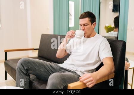 Erfolgreicher männlicher Kunde, der von einer Tasse Kaffee schlürft, während er komfortabel in der modernen und einladenden Lobby der medizinischen Klinik wartet. Stockfoto