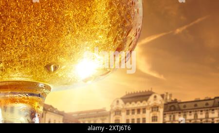 Goldenes Bier im Glas, funkelnd mit Blasen und Tropfen, mit reflektierendem Sonnenlicht und historischen Gebäuden im Hintergrund. Stockfoto