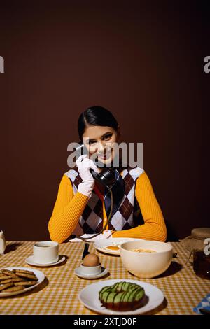 Stilvolle Frau in gelbem Pullover am Tisch, die während des Frühstücks am Drehtelefon spricht. Stockfoto
