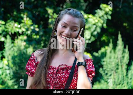 Eine schöne rothaarige Frau lächelt und redet auf ihrem Telefon, während sie durch einen sonnigen Garten spaziert und den Tag genießt. Ihre positive Energie und ihr Selbstvertrauen Stockfoto