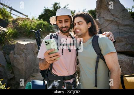 Ein junges schwules Paar macht eine Sommerwanderung und schaut zusammen auf ihr Handy, während es Rucksäcke trägt. Stockfoto