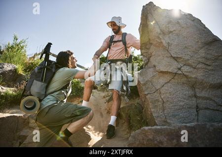 Ein junges schwules Paar wandert zusammen in der Wildnis an einem sonnigen Sommertag, ein Mann hilft dem anderen auf einem felsigen Pfad. Stockfoto