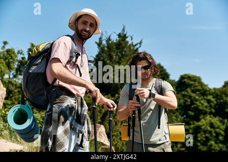 Zwei junge Männer, ein schwules Paar, wandern zusammen durch einen üppigen Sommerwald. Beide tragen Rucksäcke und halten Wanderstöcke. Stockfoto