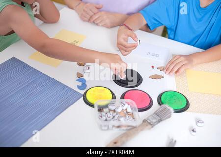 Schreibtisch mit Bastelutensilien und Kinderhänden, die Tinte zum Stempeln mit Briefmarken aufnehmen. Stockfoto