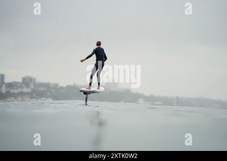 Ein junger Mann in einem Neoprenanzug reitet im Meer und fliegt über dem Wasser auf einem elektrisch betriebenen Tragflächenbrett, einer Hybridfolie mit Eoil-Folienantrieb Stockfoto