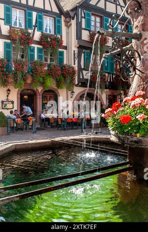 KAYSERSBERG ALSCACE Springbrunnen mit Blumendecken auf dem zentralen Platz mit Restaurant Hotel du Chateau dahinter im Elsass Kaysersberg Frankreich Stockfoto