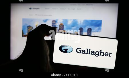 Person, die ein Mobiltelefon mit dem Logo der amerikanischen Versicherungsgesellschaft Arthur J. Gallagher Co. (AJG) vor der Webseite hält. Schwerpunkt auf dem Display des Telefons. Stockfoto