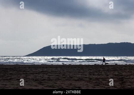 Die Silhouette eines Mannes, der seinen Hund am späten Nachmittag entlang des Strandes in Three Cliffs Bay in Wales führt. Stockfoto