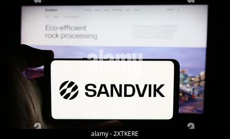 Person, die ein Mobiltelefon mit dem Logo des schwedischen Ingenieurunternehmens Sandvik ab vor der Webseite hält. Schwerpunkt auf dem Display des Telefons. Stockfoto