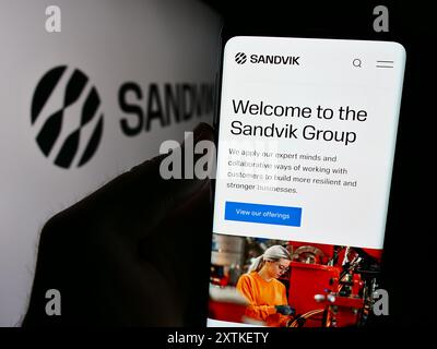 Person, die ein Handy hält und die Webseite des schwedischen Ingenieurunternehmens Sandvik ab vor dem Firmenlogo zeigt. Konzentrieren Sie sich auf die Mitte des Telefondisplays. Stockfoto