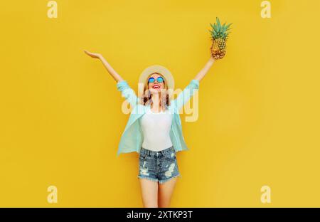 Glückliche fröhliche Frau hebt ihre Hände mit Ananas hoch, viel Spaß im Sommerhut auf gelbem Hintergrund Stockfoto