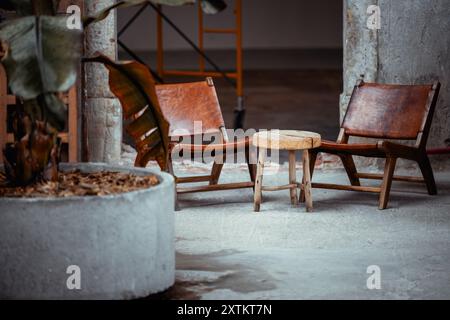 Zwei rustikale Holzstühle mit Ledersitzen und einem kleinen Holztisch in einem unvollendeten Raum. Großer Beton-Pflanzgefäß mit einer Bananenpflanze im for Stockfoto