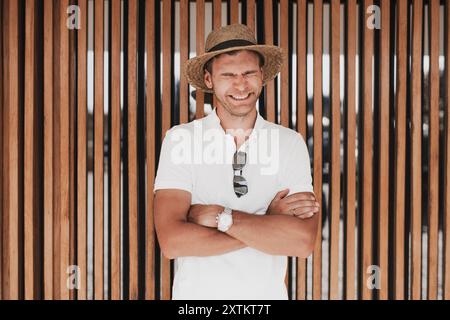 Ein lachender Mann mit Strohhut und weißem Poloshirt steht mit überkreuzten Armen vor einer hölzernen Lattenwand. Das helle, natürliche Licht enha Stockfoto