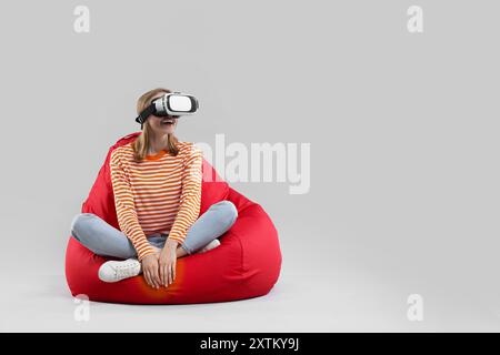 Glückliche Frau mit Virtual-Reality-Headset auf Sitzsack-Stuhl vor grauem Hintergrund, Platz für Text Stockfoto
