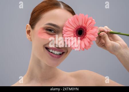 Fröhliche Frau mit klarer Haut und natürlichen Schönheitsflecken, die das Auge mit Gerbera-Blume bedecken Stockfoto