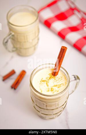 Aus nächster Nähe sehen Sie Eierlikör mit Zimt und Zimtstäbchen im nächsten Glas und eine rot-weiße Serviette. Stockfoto