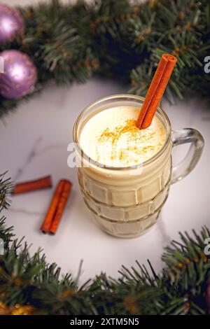 Ein Konzeptfoto eines Glases Eierpunsch mit Zimtstäbchen in einem weihnachtsriff aus nächster Nähe. Stockfoto