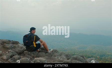 Mann, der auf Rocky Cliff Edge sitzt und unten auf Hazy Mountain View blickt, Stockfoto