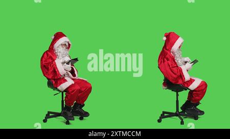 Der moderne weihnachtsmann arbeitet während der weihnachtsfeier auf einem digitalen Tablet und schaut auf der Online-Webseite, während er auf dem Stuhl sitzt. Junger Mann vor Ganzkörper-Greenscreen-Hintergrund mit Gerät. Stockfoto