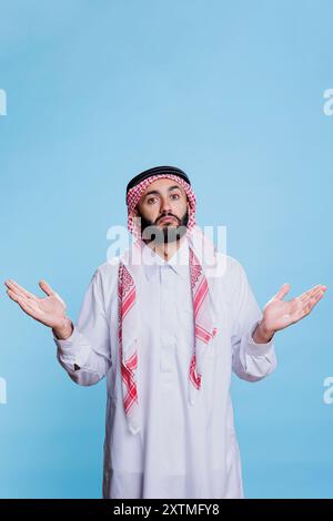 Verwirrter muslimischer Mann posiert im Studio mit offenen Armen und drückt eine zögerliche Geste aus. Arabische Person, die traditionelle Kleidung trägt, Hände spreizt und Schultern zuckt Porträt Stockfoto