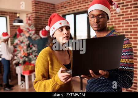 Zwei Kollegen mit weihnachtsmützen verwalten die Arbeitslast auf einem Laptop, bevor sie weihnachten im Büro feiern. Kollegen, die an einem geschmückten festlichen Arbeitsplatz stehen und einen tragbaren Computer halten Stockfoto