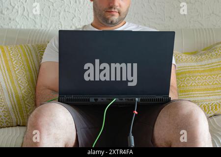 Junger Mann, der zu Hause auf dem Sofa sitzt, einen Laptop auf dem Schoß benutzt, Shorts und T-Shirt trägt Stockfoto