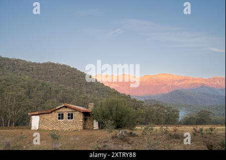 Eine Steinhütte in den Hügeln am Fuße der schneebedeckten Berge, beleuchtet vom orangefarbenen Schein einer aufgehenden Sonne mit grauem Rauch vom Lagerfeuer. Stockfoto