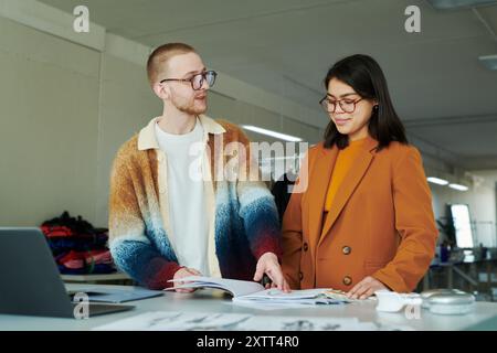 Junger männlicher Modedesigner in Brille und stilvoller Kleidung, der weibliche Kollegin ansieht, während er neue Skizzen mit ihr bespricht Stockfoto