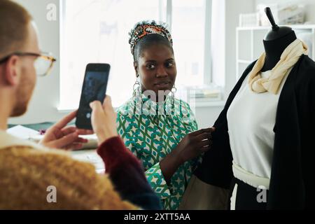 Junge afroamerikanische Schneiderin, die auf die Kamera des Smartphones schaut, die von einem männlichen Kollegen gehalten wird und ein Foto von Modedesigner macht Stockfoto