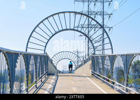 Los Angeles, Kalifornien - 21. April 2024: Radfahrer fahren auf einer Fußgängerbrücke über den Los Angeles River mit Stromleitungen im Hintergrund. Stockfoto