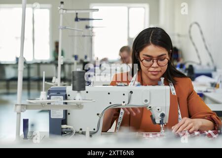 Junge Schneiderin in Brillen und Smart Casualwear Nähbekleidung am Arbeitsplatz mit Elektrogeräten in der Werkstatt Stockfoto