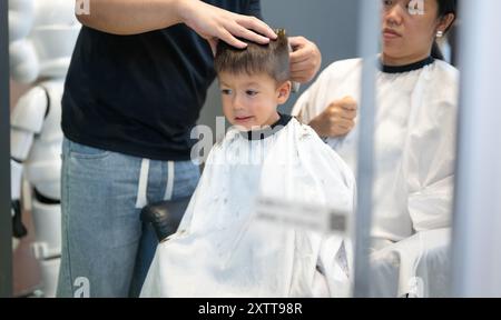 Ein kleiner Junge lächelt, als er sich von einem sanften Friseur in einem modernen Salon die Haare schneiden lässt. Die Szene fängt einen Moment des Glücks und Vertrauens ein, reflektiert Stockfoto